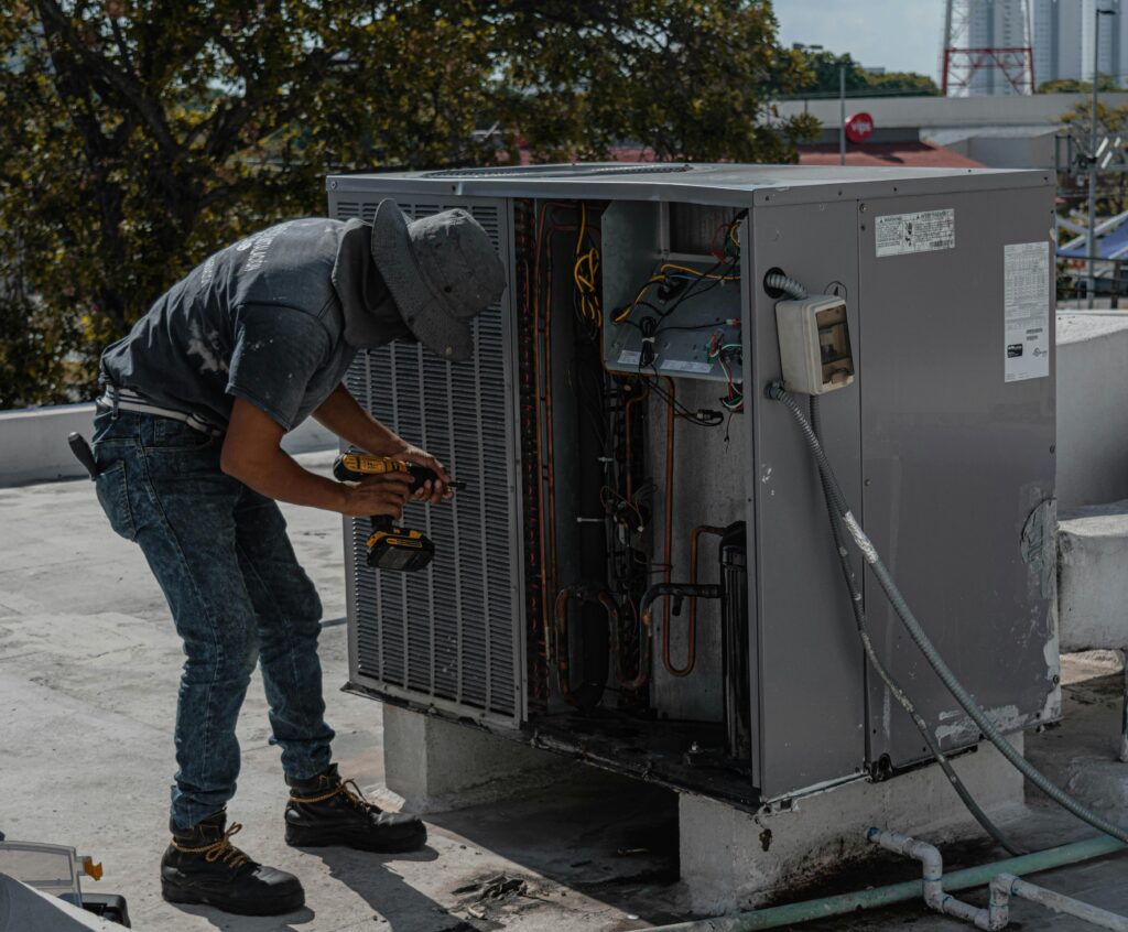 pexels-photo-5463587-5463587 A worker in a bucket hat repairs an outdoor air conditioning unit on a rooftop.